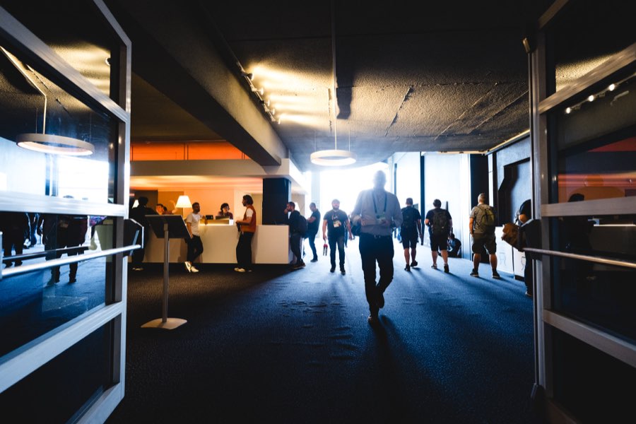 A dark carpeted corridor with pendant disc lights and an exposed concrete ceiling, an attendee silhouetted against bright daylight at the far end.