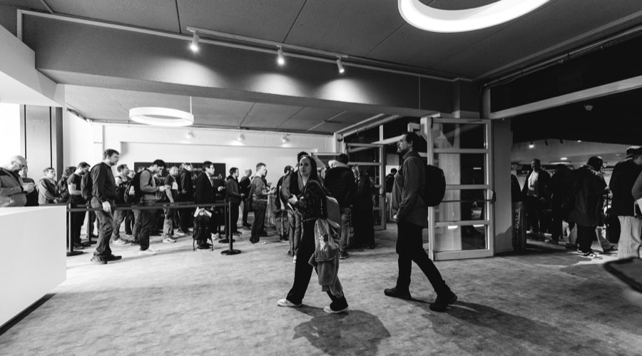 A black-and-white photograph of the foyer: attendees queueing along a long table under a circular pendant light and track spotlights.