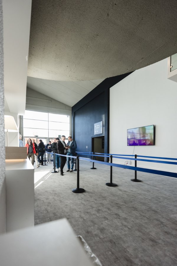 The registration area under a curved cast-concrete ceiling, with blue belt barriers across grey carpet and a wall-mounted screen beside a navy wall.