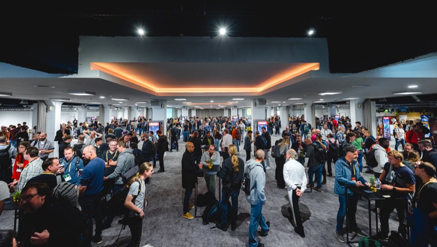 The exhibition floor packed with attendees around standing tables and booths, under a white canopy with warm cove lighting and a grid of columns.