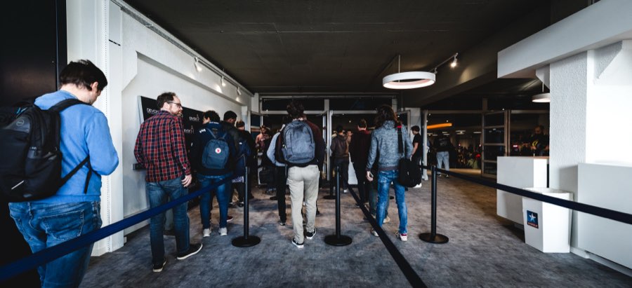 Attendees with backpacks queueing between belt barriers along a white wall towards the auditorium doors, under a dark ceiling and a disc pendant light.