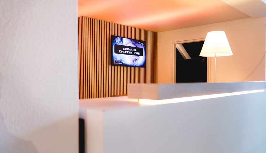 A registration desk backed by a warm slatted wood wall, with a wall-mounted screen and a table lamp.