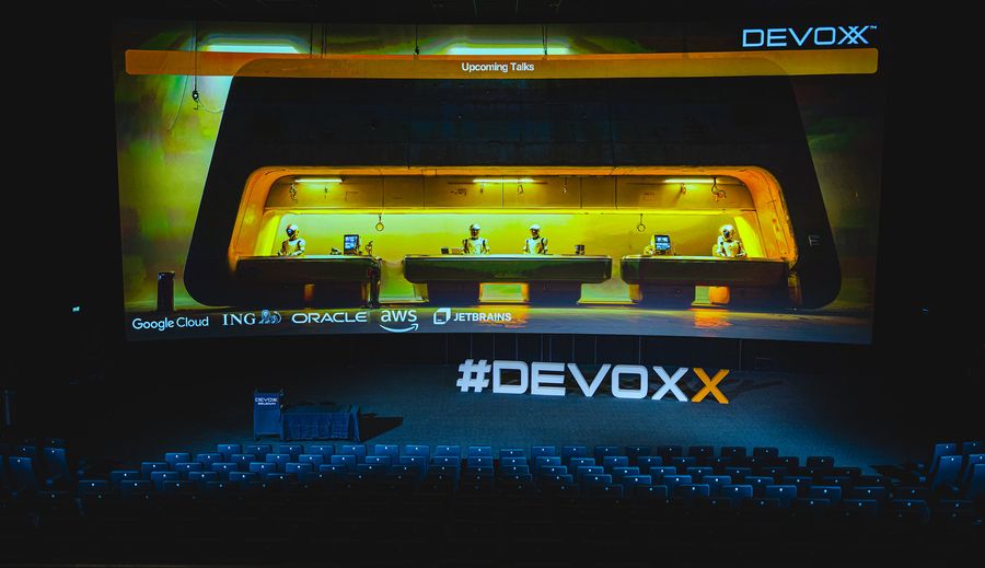 An auditorium stage lit yellow and orange, sponsor logos along the front and #DEVOXX on the screen above empty seats.
